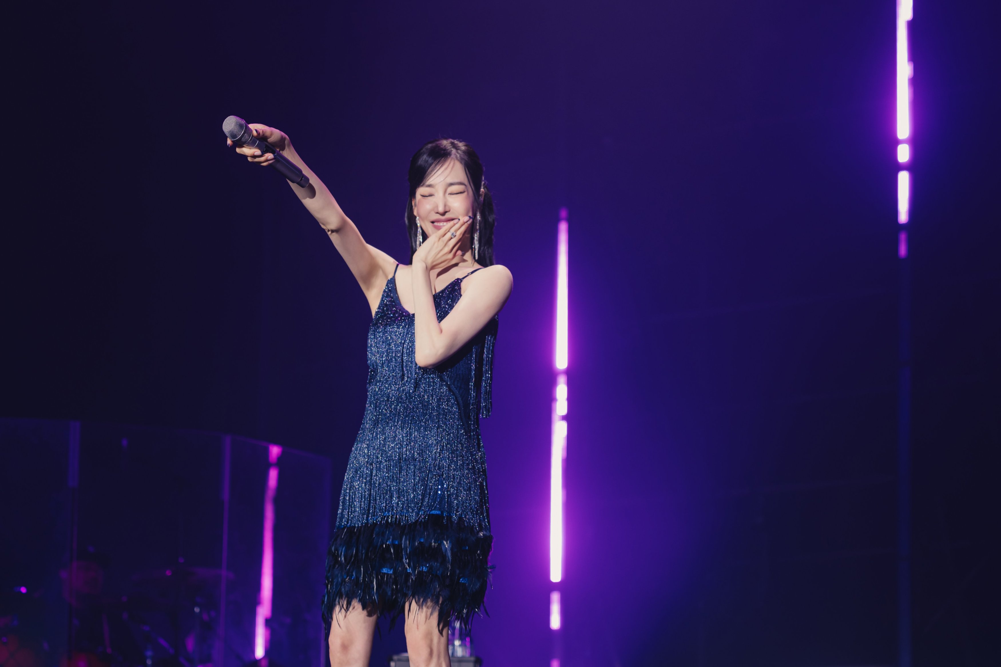 A female singer in a sparkly blue dress performs on stage, holding a microphone and smiling cheerfully with one hand on her face. Purple stage lights illuminate the background.